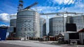 Large industrial silos are seen with metallic exteriors under a bright blue sky. The silos are interconnected with metal frameworks and are situated in an industrial area with additional smaller buildings and structures nearby. The ground is a combination of concrete and gravel, and the scene is well-lit by daylight.