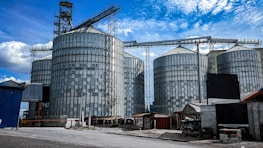 Large industrial silos are seen with metallic exteriors under a bright blue sky. The silos are interconnected with metal frameworks and are situated in an industrial area with additional smaller buildings and structures nearby. The ground is a combination of concrete and gravel, and the scene is well-lit by daylight.