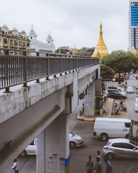 A cityscape featuring a prominent golden stupa in the background, flanked by traditional colonial-style buildings and a modern office tower. A concrete bridge crosses over a busy street below, with several vehicles and a few pedestrians visible, including people walking and riding motorcycles.