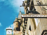 A building facade with a decorative sign reading 'The People's Story' showing a figure holding a scroll. The structure has an old-fashioned clock tower with spires in the background, and the sky is bright blue.