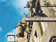 A building facade with a decorative sign reading 'The People's Story' showing a figure holding a scroll. The structure has an old-fashioned clock tower with spires in the background, and the sky is bright blue.