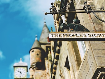 A building facade with a decorative sign reading 'The People's Story' showing a figure holding a scroll. The structure has an old-fashioned clock tower with spires in the background, and the sky is bright blue.