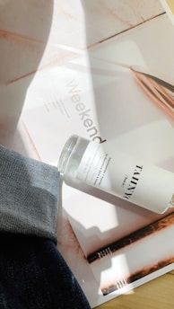 A bottle labeled with 'TAMNYC' rests on a magazine page with the word 'Weekend' partially visible. There is a hint of denim fabric draped across the lower left corner, and sunlight creates shadows on the surface.