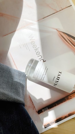A bottle labeled with 'TAMNYC' rests on a magazine page with the word 'Weekend' partially visible. There is a hint of denim fabric draped across the lower left corner, and sunlight creates shadows on the surface.