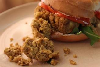 A close-up of a burger made with a crumbly, textured patty likely made from chickpeas or beans. The burger is topped with slices of tomato and green lettuce, placed in a soft, lightly browned bun. Crumbs from the patty are scattered on the plate, adding a rustic look.