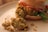 A close-up of a burger made with a crumbly, textured patty likely made from chickpeas or beans. The burger is topped with slices of tomato and green lettuce, placed in a soft, lightly browned bun. Crumbs from the patty are scattered on the plate, adding a rustic look.