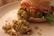 A close-up of a burger made with a crumbly, textured patty likely made from chickpeas or beans. The burger is topped with slices of tomato and green lettuce, placed in a soft, lightly browned bun. Crumbs from the patty are scattered on the plate, adding a rustic look.