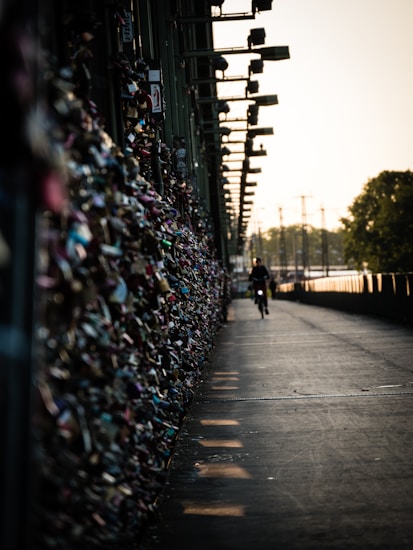 A pathway is bordered by a wall covered in numerous padlocks, extending into the distance. A solitary cyclist rides along the concrete path, with silhouetted trees and a setting sun in the background creating a warm glow.