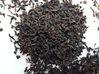 A pile of loose, dark brown to nearly black tea leaves scattered on a white surface. The leaves appear curled and twisted, with various sizes and textures.