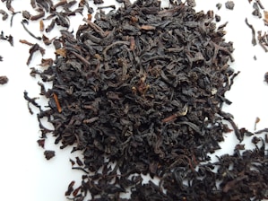 A pile of loose, dark brown to nearly black tea leaves scattered on a white surface. The leaves appear curled and twisted, with various sizes and textures.