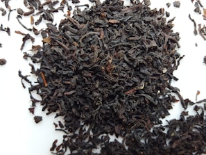 A pile of loose, dark brown to nearly black tea leaves scattered on a white surface. The leaves appear curled and twisted, with various sizes and textures.