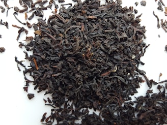 A pile of loose, dark brown to nearly black tea leaves scattered on a white surface. The leaves appear curled and twisted, with various sizes and textures.