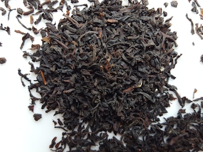 A pile of loose, dark brown to nearly black tea leaves scattered on a white surface. The leaves appear curled and twisted, with various sizes and textures.