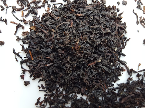 A pile of loose, dark brown to nearly black tea leaves scattered on a white surface. The leaves appear curled and twisted, with various sizes and textures.
