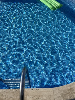 Clear blue water ripples in a swimming pool with a shiny metal ladder at the pool's edge and a green inflatable float on the water surface.