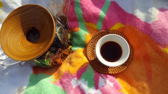 A wooden pour-over coffee maker with a patterned glass bottom sits on a colorful towel. Beside it, there is a small white cup filled with dark coffee, resting on a woven coaster with an orange hue. The towel features vibrant colors including shades of pink, orange, green, and yellow, resembling a floral pattern.