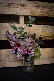 A rustic bouquet of wildflowers arranged in a transparent mason jar sits against a backdrop of stacked wooden crates. The arrangement includes purple coneflowers, various green leaves, and other wild blossoms. The crates have text stamped on them and exhibit a weathered texture.