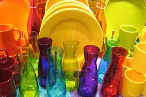 A collection of colorful glassware and tableware featuring various translucent bottles, mugs, and plates. The vibrant colors include red, yellow, blue, green, and purple, arranged closely together, creating a visually striking display.