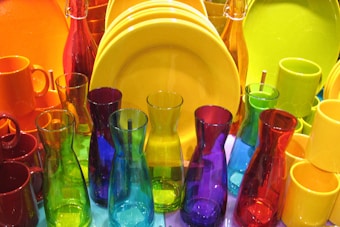 A collection of colorful glassware and tableware featuring various translucent bottles, mugs, and plates. The vibrant colors include red, yellow, blue, green, and purple, arranged closely together, creating a visually striking display.