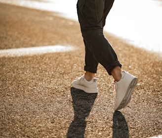 person in black pants and white sneakers walking on brown asphalt road during daytime