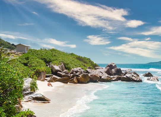 A serene tropical beach scene featuring turquoise waters gently lapping against a white sandy shore. Large, smooth boulders are scattered along the edge of the sand, surrounded by lush green vegetation. A small building is visible among the greenery. The sky is clear with a few scattered clouds, creating a peaceful and inviting atmosphere.