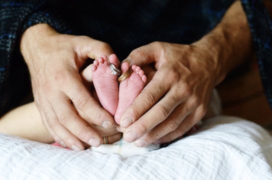 A close-up view of an adult's hands gently holding a newborn baby's feet, with wedding rings placed on the baby's toes. The soft lighting enhances the tenderness of the scene.