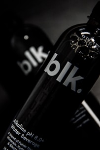 A close-up view of a black bottle with the text 'blk.' on the front. The bottle appears to contain an alkaline water beverage with a pH of 8.0 or higher. The background is dark, focusing attention on the sleek design of the bottle, with some reflections visible on its surface.