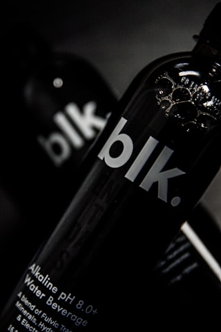 A close-up view of a black bottle with the text 'blk.' on the front. The bottle appears to contain an alkaline water beverage with a pH of 8.0 or higher. The background is dark, focusing attention on the sleek design of the bottle, with some reflections visible on its surface.