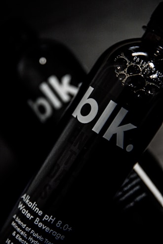 A close-up view of a black bottle with the text 'blk.' on the front. The bottle appears to contain an alkaline water beverage with a pH of 8.0 or higher. The background is dark, focusing attention on the sleek design of the bottle, with some reflections visible on its surface.