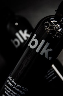 A close-up view of a black bottle with the text 'blk.' on the front. The bottle appears to contain an alkaline water beverage with a pH of 8.0 or higher. The background is dark, focusing attention on the sleek design of the bottle, with some reflections visible on its surface.