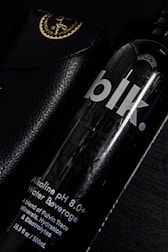 A black bottle labeled 'blk.' with text indicating it is an alkaline pH 8.0+ water beverage. The bottle is partially overlapping a black leather case with golden writing and an emblem at the corner. The setting is predominantly dark, with highlights reflecting off the glossy surfaces.
