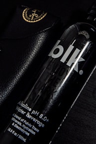 A black bottle labeled 'blk.' with text indicating it is an alkaline pH 8.0+ water beverage. The bottle is partially overlapping a black leather case with golden writing and an emblem at the corner. The setting is predominantly dark, with highlights reflecting off the glossy surfaces.