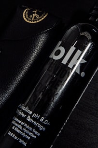 A black bottle labeled 'blk.' with text indicating it is an alkaline pH 8.0+ water beverage. The bottle is partially overlapping a black leather case with golden writing and an emblem at the corner. The setting is predominantly dark, with highlights reflecting off the glossy surfaces.
