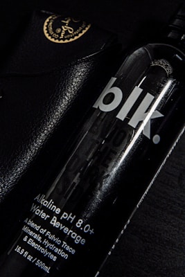 A black bottle labeled 'blk.' with text indicating it is an alkaline pH 8.0+ water beverage. The bottle is partially overlapping a black leather case with golden writing and an emblem at the corner. The setting is predominantly dark, with highlights reflecting off the glossy surfaces.