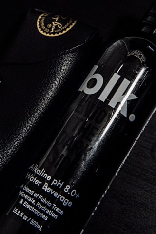 A black bottle labeled 'blk.' with text indicating it is an alkaline pH 8.0+ water beverage. The bottle is partially overlapping a black leather case with golden writing and an emblem at the corner. The setting is predominantly dark, with highlights reflecting off the glossy surfaces.