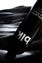 A black water bottle with the label 'blk' in white font is placed against a textured, dark background. The background features a soft, wave-like pattern with a gradient of black and gray shades. The image has a sleek, modern appearance due to the contrast between the dark bottle and the lighter background.