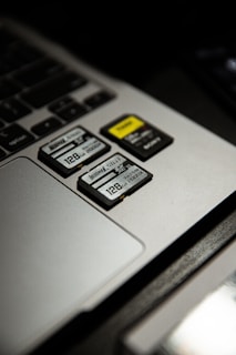 Three SD cards are placed on a laptop's touchpad. One of the cards has a yellow label, and all cards display a storage capacity of 128 GB. The laptop's keyboard is partially visible beside the touchpad.