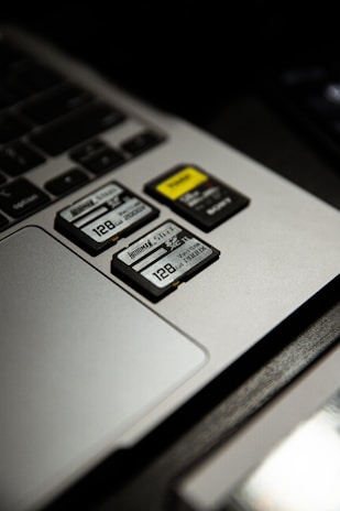 Three SD cards are placed on a laptop's touchpad. One of the cards has a yellow label, and all cards display a storage capacity of 128 GB. The laptop's keyboard is partially visible beside the touchpad.