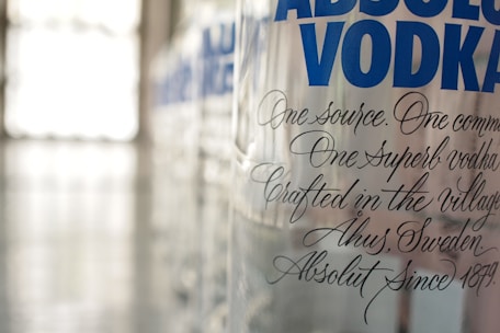 A close-up view of a clear glass bottle with the words 'ABSOLUT VODKA' prominently displayed in bold blue letters. The bottle features elegant, scripted black text detailing its craftsmanship, origin in Åhus, Sweden, and the year 1879. The background is softly blurred, giving emphasis to the bottle.
