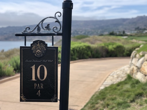 A metal pole with an ornate design at the top holds a sign that reads 'Trump National Golf Club 10 PAR 4'. The background includes a curved pathway lined with greenery and stone, leading to a golf course with a scenic, hilly landscape and a cloudy sky.
