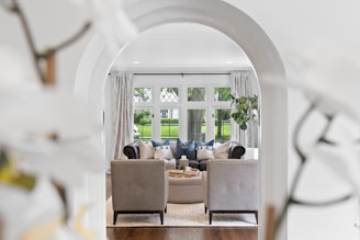 A beautifully arranged living room viewed through an arched doorway, with two armchairs facing a plush couch. The room is elegantly decorated with cushions and a central coffee table that houses candles and small plants. Large windows with lattice patterns offer a view of a green lawn outside, and light curtains frame the scene. A potted plant adds a touch of greenery to the interior ambiance.