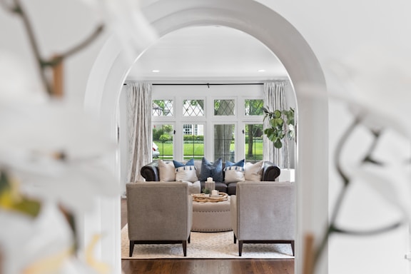 A beautifully arranged living room viewed through an arched doorway, with two armchairs facing a plush couch. The room is elegantly decorated with cushions and a central coffee table that houses candles and small plants. Large windows with lattice patterns offer a view of a green lawn outside, and light curtains frame the scene. A potted plant adds a touch of greenery to the interior ambiance.