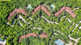 Aerial view of a zig-zag structured complex surrounded by dense greenery, with two visible swimming pools and a tennis court. The buildings are brown-roofed, blending with the environment. Paths and roads weave through the trees, providing access to the various facilities.