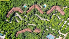 Aerial view of a zig-zag structured complex surrounded by dense greenery, with two visible swimming pools and a tennis court. The buildings are brown-roofed, blending with the environment. Paths and roads weave through the trees, providing access to the various facilities.