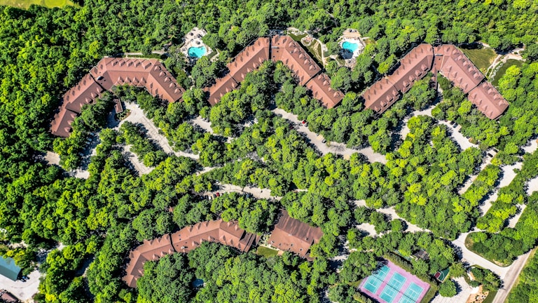 Aerial view of a zig-zag structured complex surrounded by dense greenery, with two visible swimming pools and a tennis court. The buildings are brown-roofed, blending with the environment. Paths and roads weave through the trees, providing access to the various facilities.