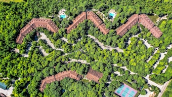 Aerial view of a zig-zag structured complex surrounded by dense greenery, with two visible swimming pools and a tennis court. The buildings are brown-roofed, blending with the environment. Paths and roads weave through the trees, providing access to the various facilities.