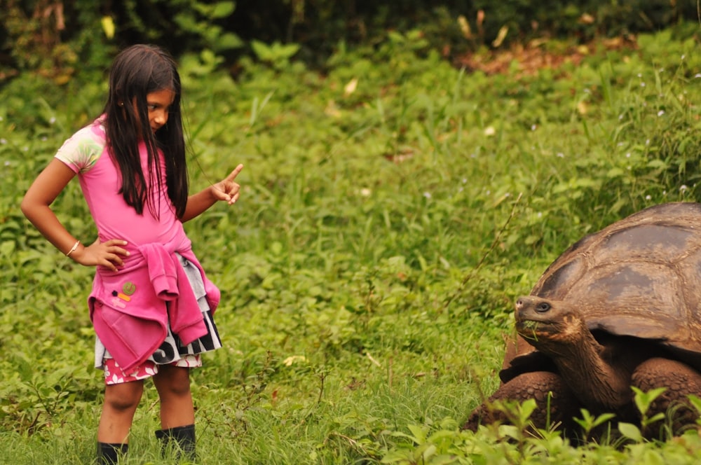 낮 동안 푸른 잔디에 갈색 동물 옆에 서 있는 분홍색 드레스를 입은 소녀