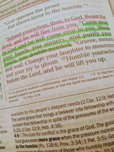 A close-up of a Bible page with highlighted text in pink and green. The main verse visible is 'Submit yourselves, then, to God. Resist the devil, and he will flee from you.' The text is written in a serif font, and there are footnotes and references included at the bottom of the page. The highlighted sections emphasize phrases about devotion and coming near to God.