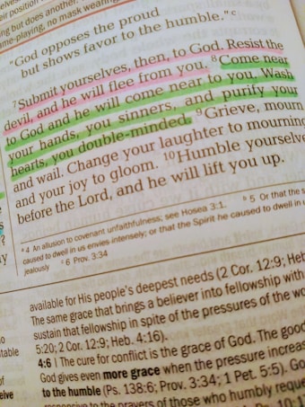 A close-up of a Bible page with highlighted text in pink and green. The main verse visible is 'Submit yourselves, then, to God. Resist the devil, and he will flee from you.' The text is written in a serif font, and there are footnotes and references included at the bottom of the page. The highlighted sections emphasize phrases about devotion and coming near to God.