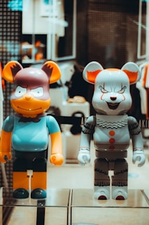 Two cartoon character figurines with large, exaggerated facial features are displayed on a shelf. The figure on the left has a humorous expression with wide eyes and is dressed in blue and orange. The figure on the right has a sinister expression with sharp teeth and is dressed in a colorful clown outfit, primarily gray with red accents.